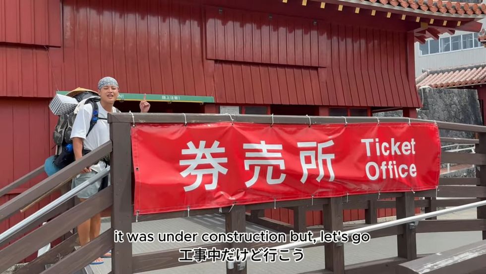 A man standing next to a banner reading "Ticket office" in English and Japanese. The onscreen text reads "it was under construction but let's go"