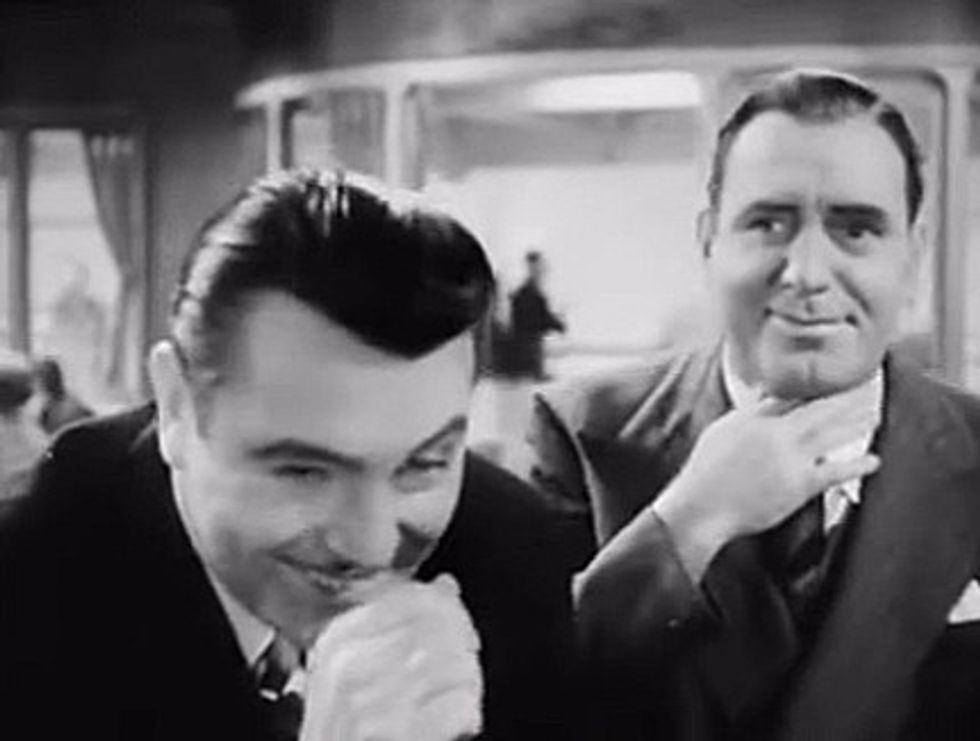 Black-and-white image of two actors, one reacting to choking on a drink.